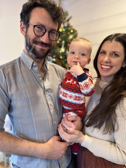 Sam Fleischer stands with wife and baby at home during the holidays. There is a decorated Christmas tree behind them.