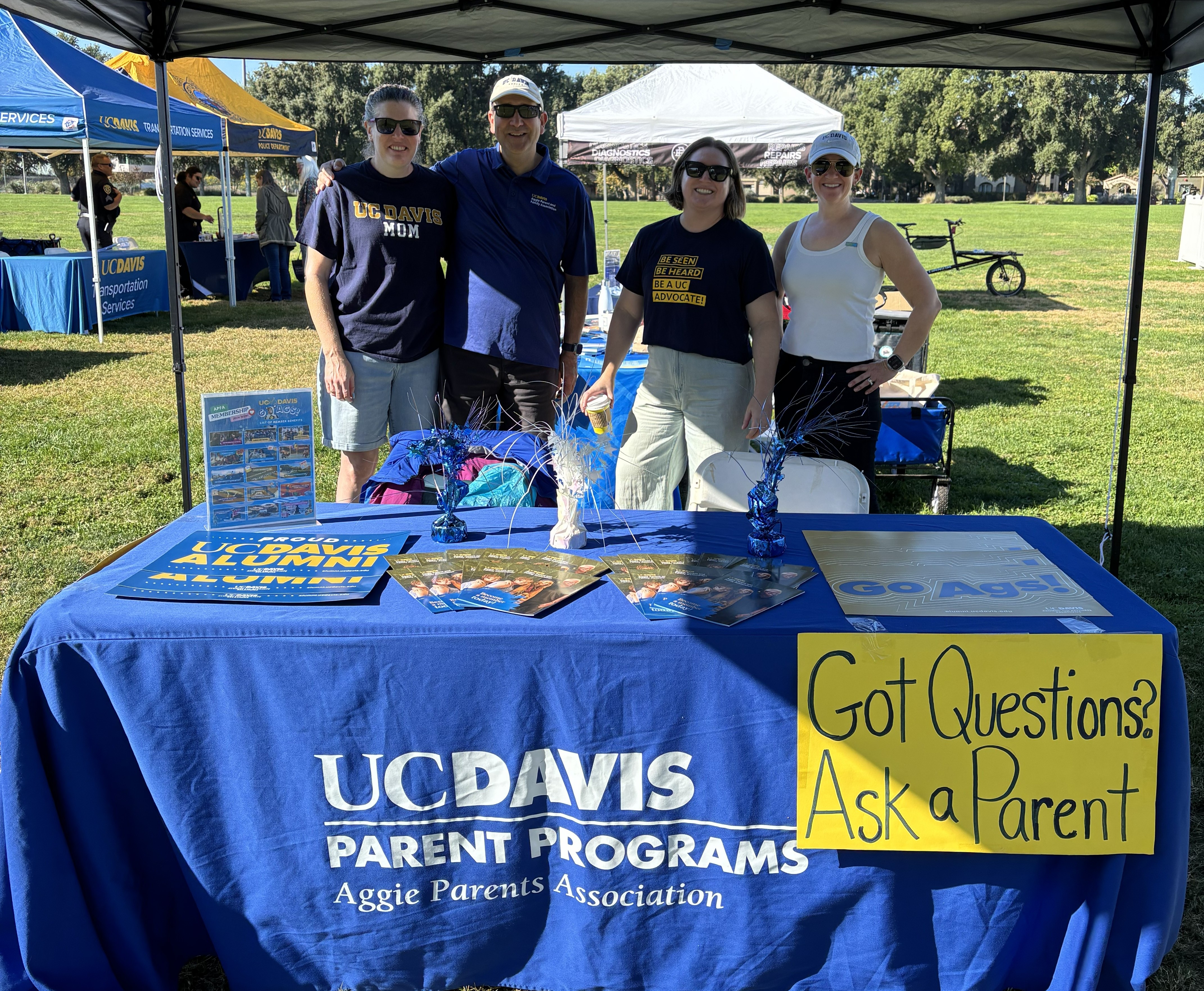 four people standing behind a table at a fair labeled "Ask a Parent"