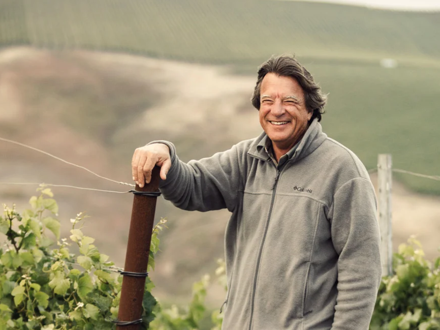 Smiling man in gray jacket leaning on post in green vineyard