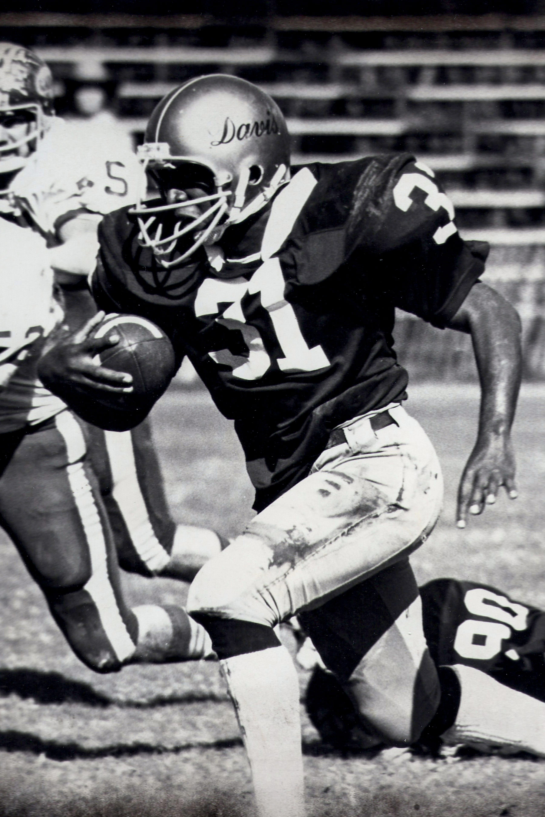 Black and white image of Ron wearing a helmet and gear while running with the football on the field.