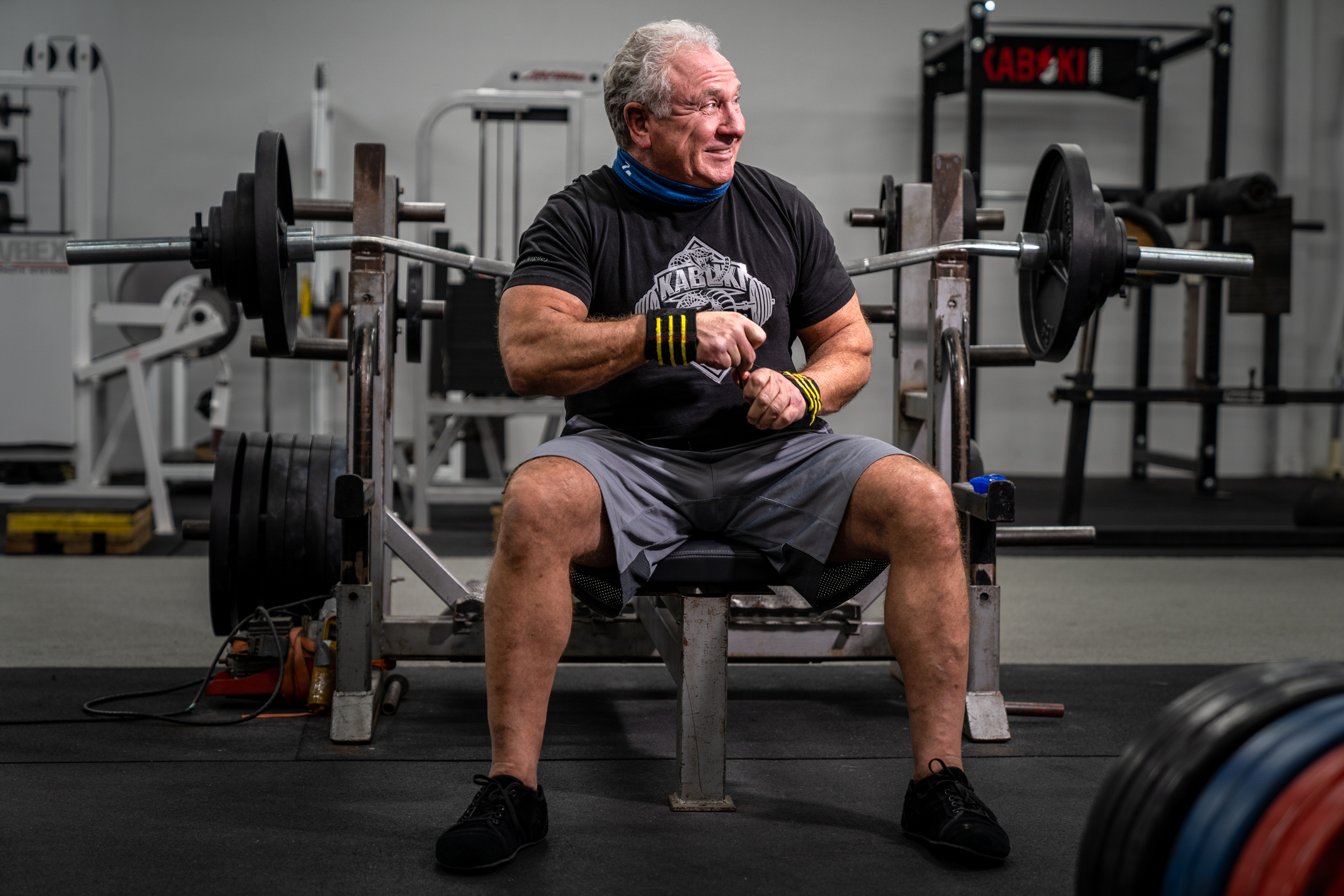 Rudy Kadlub smiles while seated on a weightlifting bench. He wears a Kabuki Strength shirt, exercise bands around his wrists, and a gaiter.
