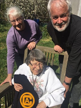 Audrey West sitting outside on a wooden chair with her daughter and son by her side, holding her UC Davis varsity-letter jacket.