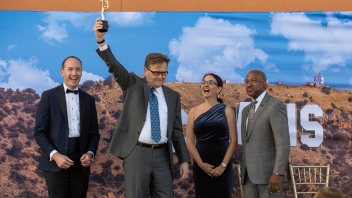 John M. Wasson ’84 lifts his golden award in the air and is flanked by former Board President Charles Melton ’08, AVC Dana Allen, and Chancellor Gary May on stage.