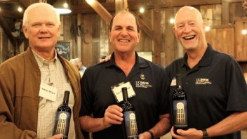Three men smiling at the camara holding wine bottles. 