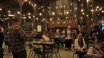 Man giving a talk about the estate and the wine in the center of the room among people sitting at tables. The photo is taken from behind him. 