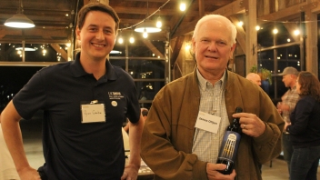 Two men smiling at the camera one is holding a wine bottle. 