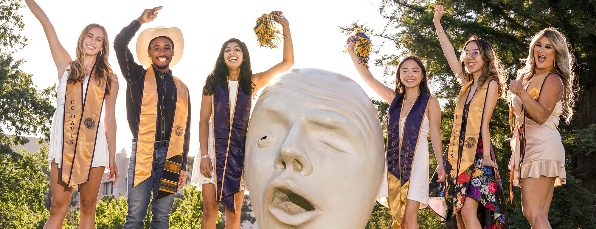 Undergraduate students in formal wear and regalia celebrating around an egg head statue.