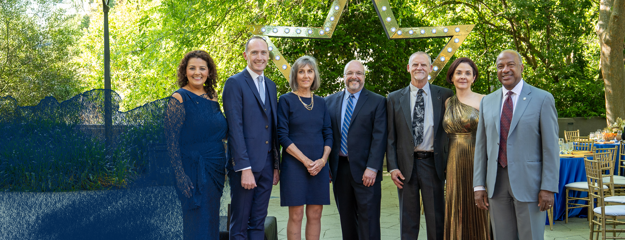 A group of UC Davis Alumni Award winners pose with Chancellor May, CAAA President Anu Johl Singh and CAAA Executive Director Dana Allen. They are posing in front of a large golden star arch on Moss Patio.