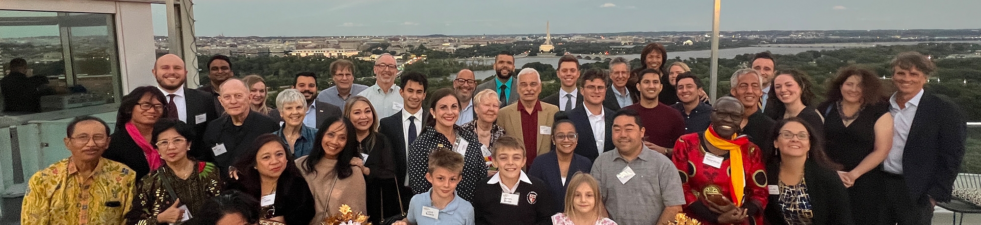 The group of attendees, association representatives, and Santi Budaya dancers wearing traditional Indonesian attire posing for a group photo on the rooftop of Top of the Town with the Potomac River and Capitol building visible in the background.