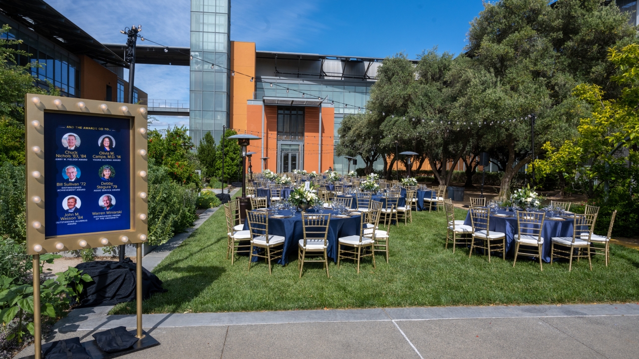 Round tables with blue linens and tablecloths, as well as gold chiavari chairs set up at the Good Life Garden for the Alumni Awards.