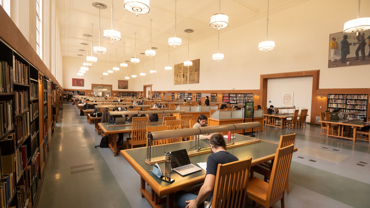 Photo of the main study room in the library, with stakes of books on the left and tables with students studying in the middle