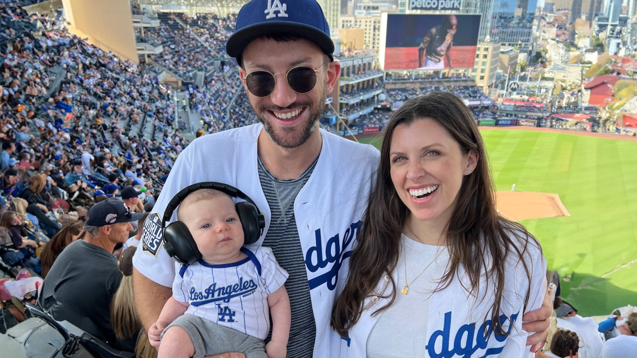 Sam Fleischer stands with wife and baby at Dodgers stadium. Each are wearing white and blue Dodgers jerseys. 