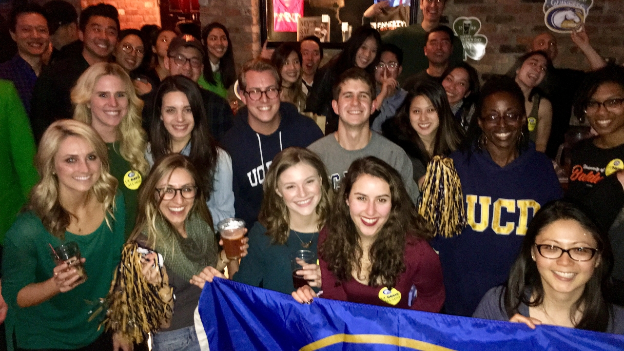 Picture includes many people grouped together, smiling at the camera, holding blue and gold pom-poms and a UCD flag