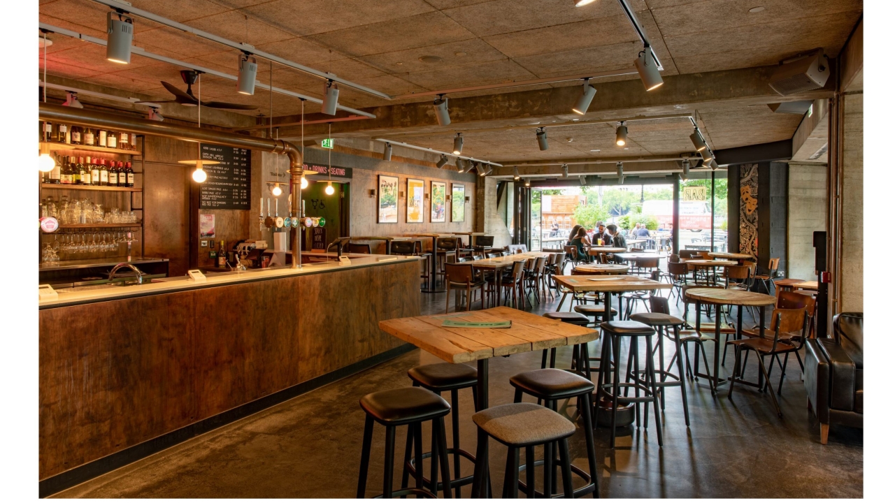 Photo of a bar with tables in the middle, and some groups of people gathered in the back of the photo