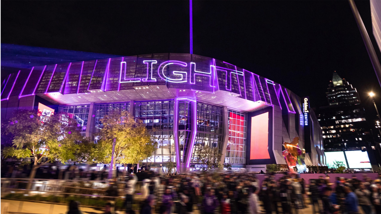 Modern arena facade lit purple at night, blurred crowd and city skyline behind