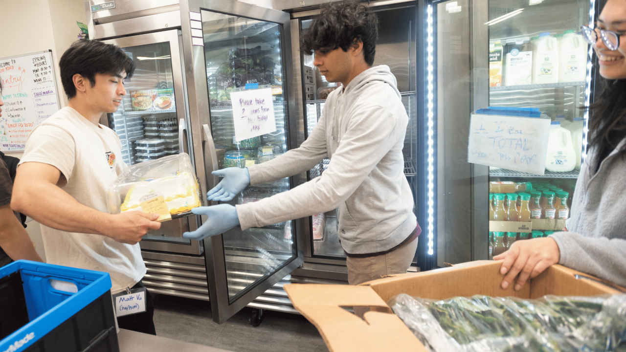 Three students in front of a fridge, the student in the middle handing food to the student on the left, and the student on the right holding a box with produce in it