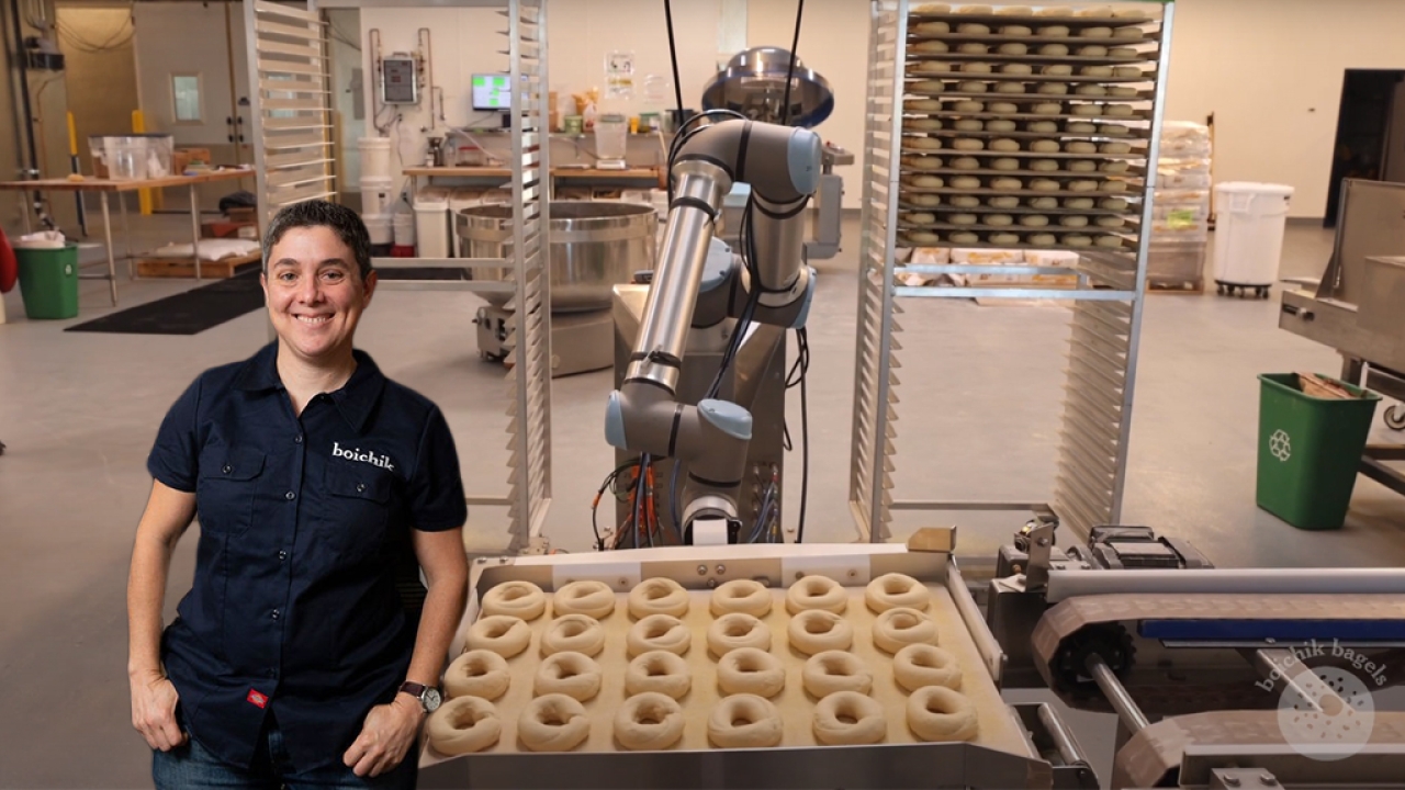 Emily Winston wearing a &#34;Boichik Bagels&#34; button-down shirt placed in front of an assembly line with a bagel-making robot and a tray of unbaked bagels.