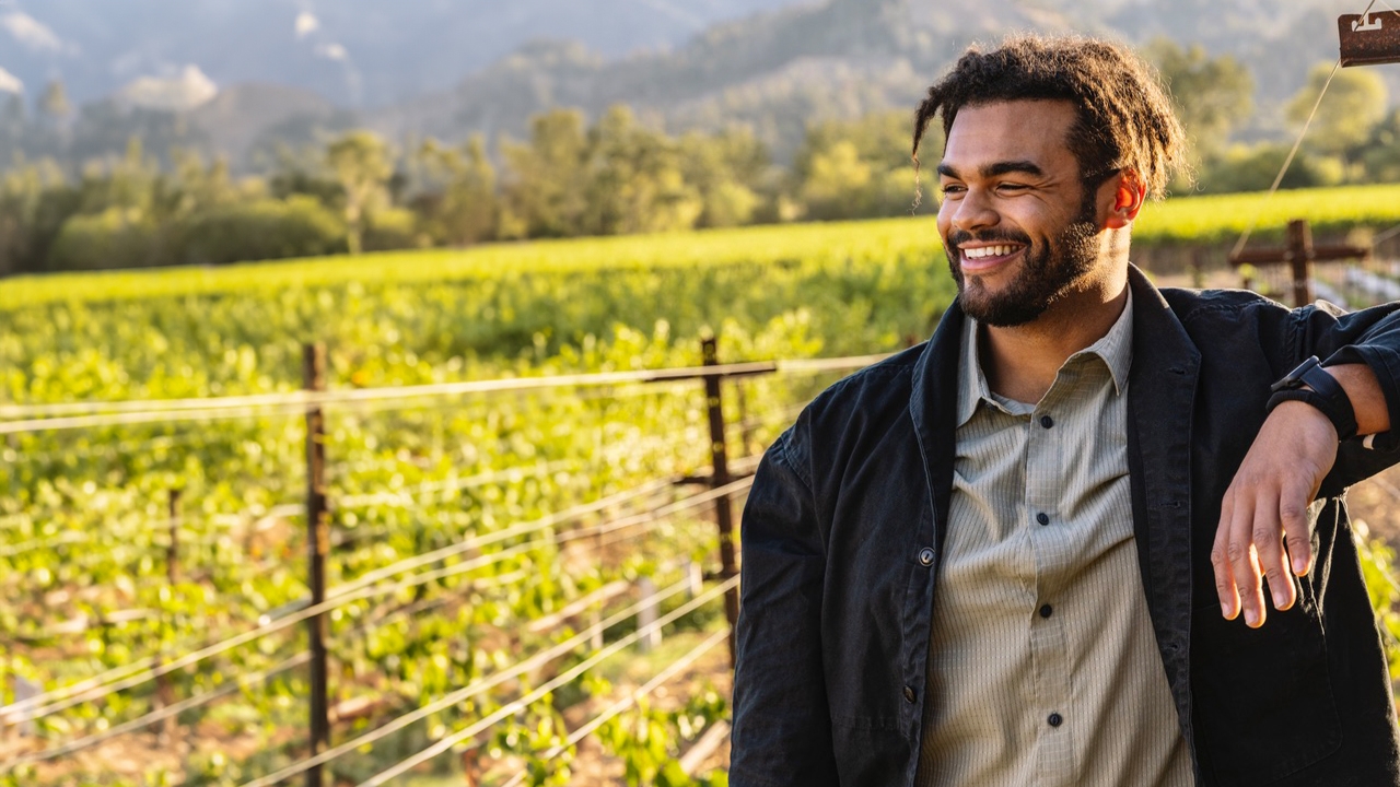 Derek poses with a black overcoat and grey button-down shirt. Vineyards and hills are visible in the background.