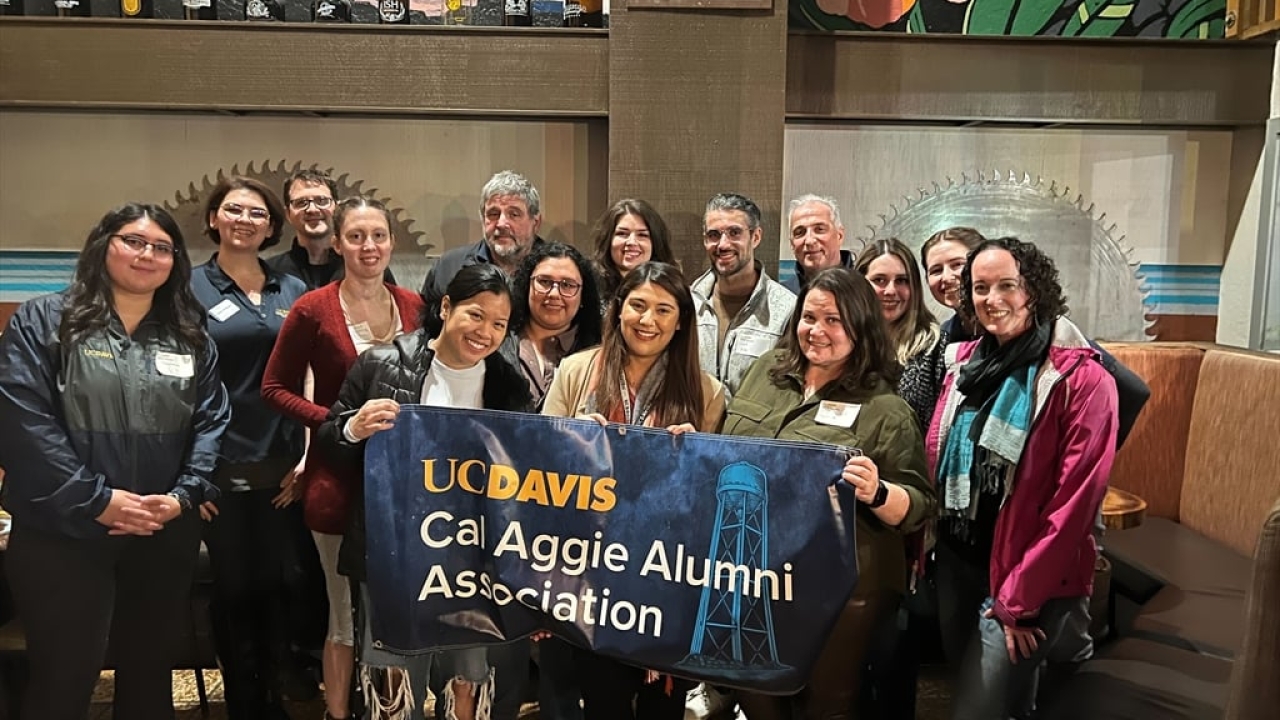 15 people facing the camera, with the 3 in front holding a banner reading, "UC Davis Cal Aggie Alumni Assocation"