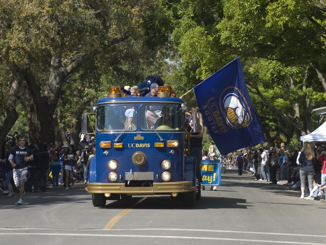 Parade at UC Davis Picnic Day. The blue UC Davis motorized float is in the center of the photo in the middle of the parade route. Someone carries the "UC Davis Aggies" flag on the parade  float.
