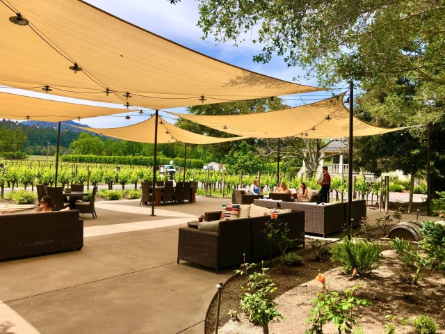 Outdoor shaded patio with tan sail canopies, wicker seating, string lights, and vineyard view.