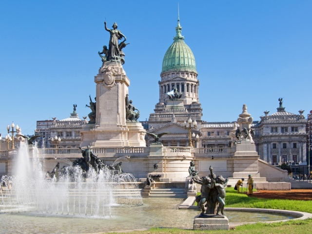 The ornate fountain in front of a historic building with a green dome and sculptures.