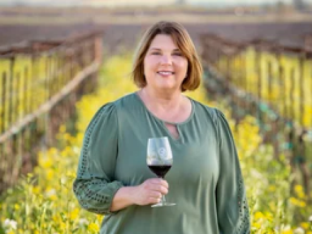 Smiling woman in green blouse holding a glass of red wine in a sunlit vineyard
