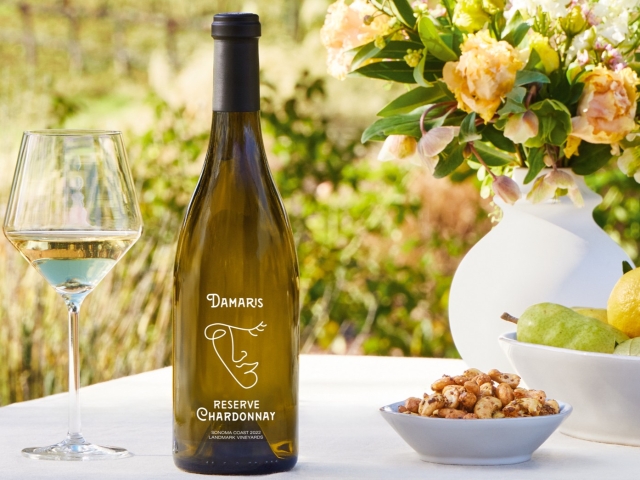 Chardonnay bottle and glass beside bowl of nuts and yellow roses on sunlit outdoor table