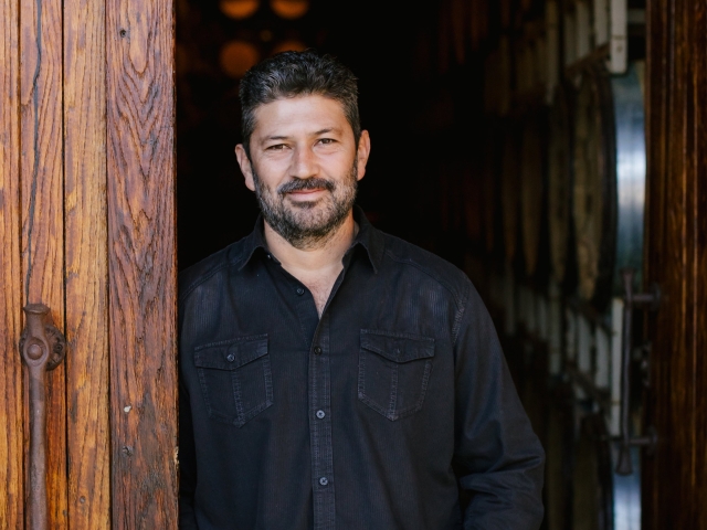 Smiling man in dark shirt by wooden post with wine barrels behind — photograph