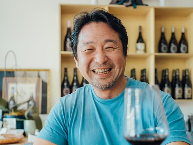 Smiling middle-aged man in blue shirt with wine glass; wine bottles on shelf behind (photo)