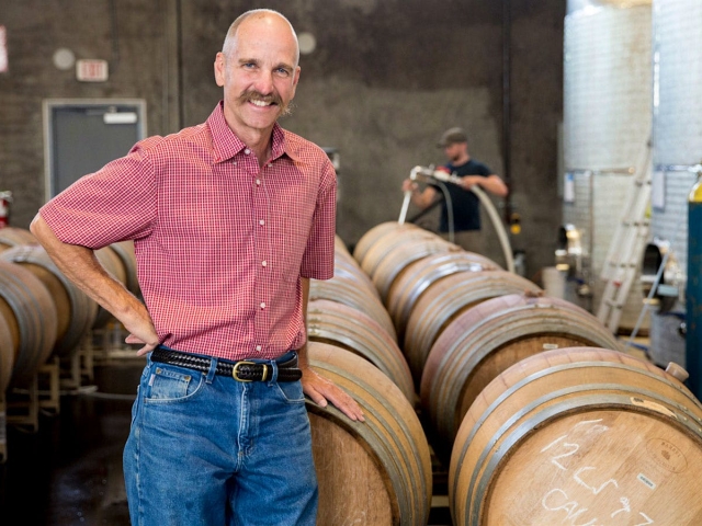 Smiling middle-aged man in red shirt standing among rows of wooden barrels in a warehouse