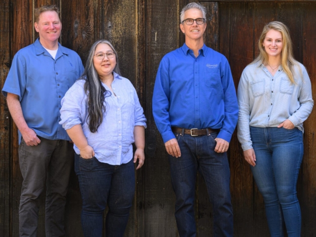 Four smiling adults in blue shirts standing against a dark wooden wall
