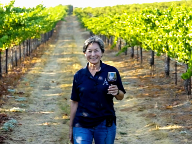 Smiling woman walking down sunlit vineyard aisle holding a wine glass and bottle.