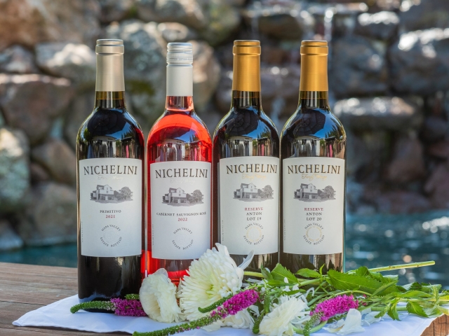 Four wine bottles on a wooden table by a pool with flowers in front