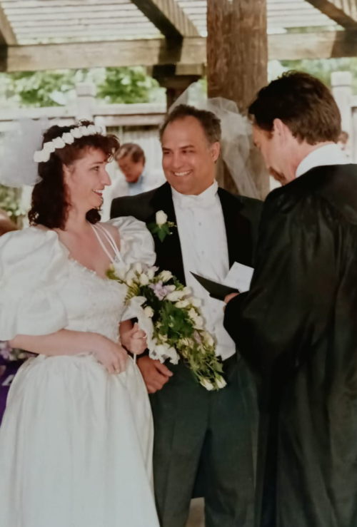 Bride and groom Donna and Tony smiling to each other at the altar in front of minister. Donna wears a white dress with a flower crown and large white bouquet of flowers.