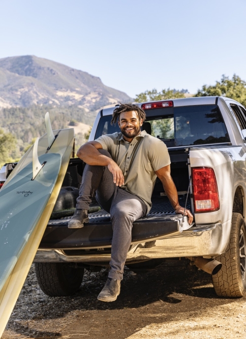 Derek poses with one leg up while sitting on the bed of a truck with a surfboard leaning on the truck. Hills are visible in the background.