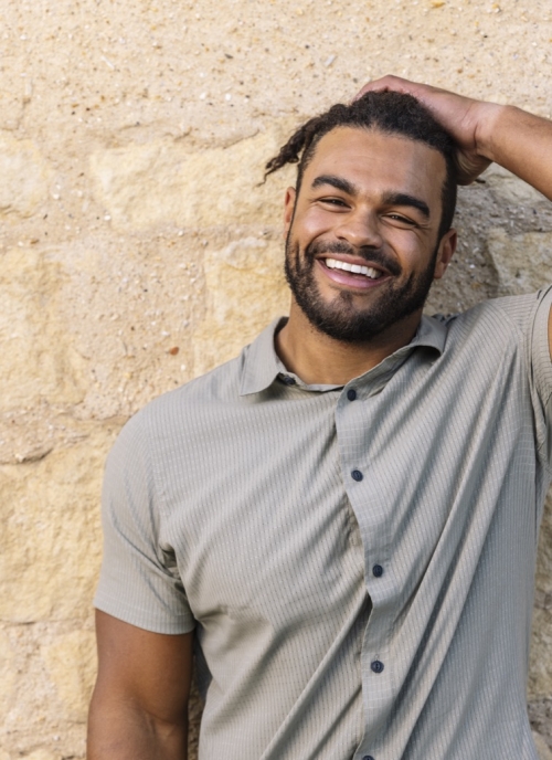 Derek poses in a collared button-down shirt with one hand on his head in front of a sandstone wall.