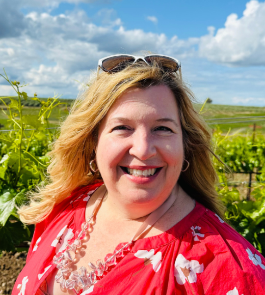 Ginger Welsh wearing sunglasses on her head and a red top with white flowers in front of a vineyard with blue skies.