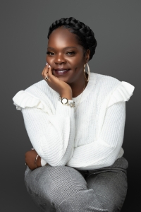 Professional headshot of Bland sitting cross-legged, hand under chin and smiling and looking at the camera