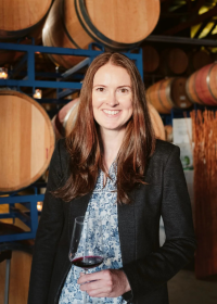 Smiling woman in blazer holding a glass of red wine in a barrel room