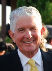 Older male poses for a photo outdoors wearing a white button-down, gold tie, and dark blazer.