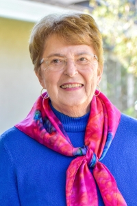 Older female poses outdoors wearing a blue sweater, fuchsia scarf, glasses, and gold hoop earrings.