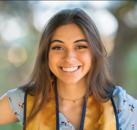 Ashlee Peterson wearing a floral print dress and a blue and gold grad stole smiling in front of a blurry bokeh background.