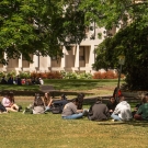 A group of students sitting on the grass in front of trees and a building in the background