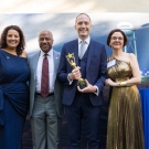 Four people on stage at the awards ceremony. Charles Melton in the middle holds the gold statue.
