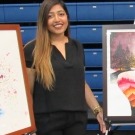 A woman with long brown hair, wearing a black outfit, stands smiling in front of three framed paintings displayed on easels in an indoor exhibition space. The artwork on the left features an abstract composition with splashes of color and floral elements. The middle piece portrays a surreal landscape with layered, colorful hills inside a floating shape against a night sky with trees. The artwork on the right consists of a dark blue abstract form with splattered paint on a white background. The setting inclu