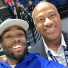 Two people pose for a selfie at a UC Davis sporting event. One person is wearing a white ballcap and blue sweatshirt and the other is wearing a gray suit with a blue and yellow checkered shirt.