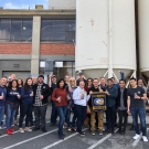 Tour group of 21 people standing outside a factory cheerings to the camera with beer glasses, with two people holding a UC Davis Aggies banner