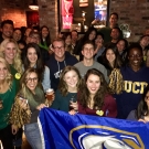 Picture includes many people grouped together, smiling at the camera, holding blue and gold pom-poms and a UCD flag