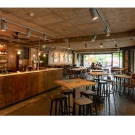 Photo of a bar with tables in the middle, and some groups of people gathered in the back of the photo
